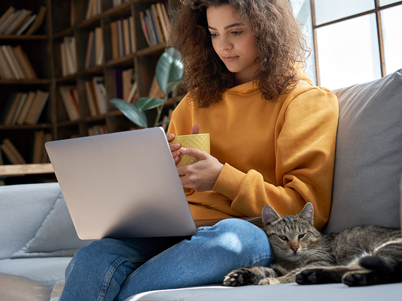 attractive young woman sitting on the couch with laptop open and a tabby kitty snuggling beside her.